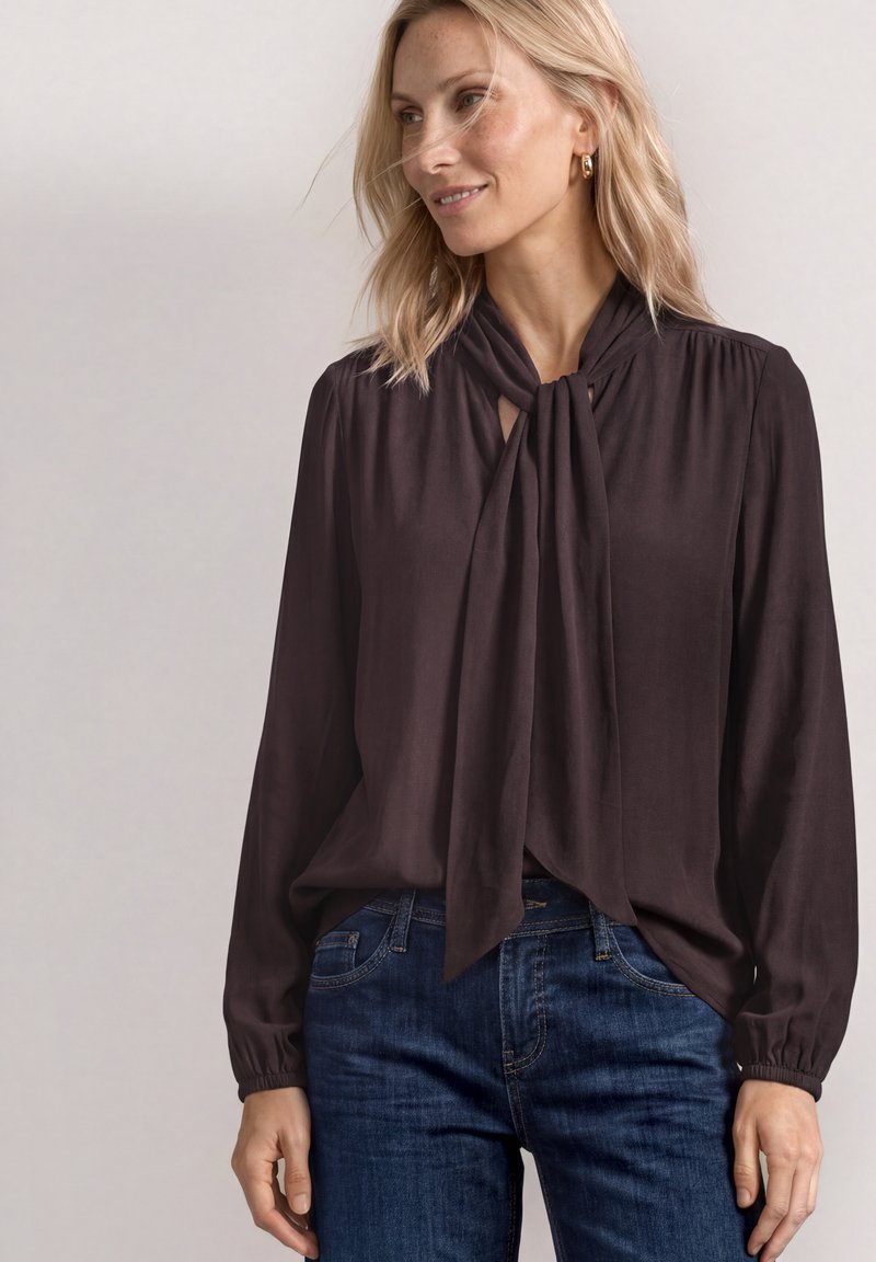 Femme blonde portant un chemisier noir à manches longues avec un col noué et un jean bleu, souriante, sur un fond neutre uni.
