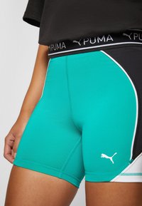 Zbliżenie osoby noszącej szmaragdowe i czarne szorty sportowe Puma z białym logo oraz czarnym pasem z brandingiem Puma.
