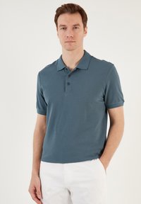 Paita polo dalam warna teal gelap, lengan pendek, dengan plaket tiga butang dan kain bertekstur; dipadukan dengan celana putih.