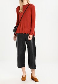 Roter Strickpullover mit gewelltem Saum, schwarze Leder-Culottes und braune Wildleder-Stiefeletten mit Blockabsatz; trägt eine kleine schwarze Umhängetasche.