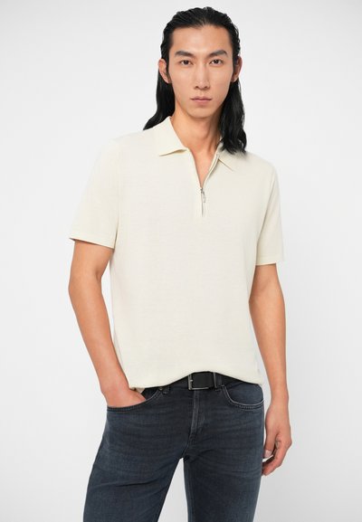 Άνδρας με μακριά μαύρα μαλλιά, φορώντας κρέμ πόλο μπλούζα με κοντά μανίκια και φερμουάρ, μέσα σε σκούρο μπλε τζιν με μαύρη ζώνη, το χέρι του στην τσέπη.