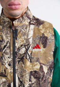 Camo-mønstrede zip-up jakke i brun og grøn; har et adidas-logo i rød. Struktureret stof med høj krave og sort lynlås.