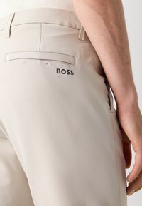 Calças bege com um tecido suave e leve. Apresenta um bolso traseiro e o logótipo 'BOSS' impresso a preto perto da cintura.