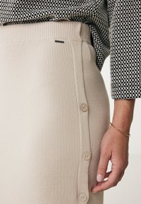 Beige ribbad stickad kjol med hög midja och sidoknappdetalj, parat med en kontrasterande svart och vit mönstrad topp. Slät textur.