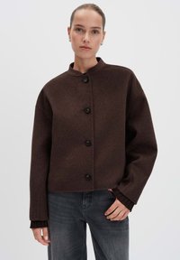 Vrouw in een donkerbruine gewatteerde wollen jas met een ronde kraag en donkergrijze jeans, staand tegen een effen lichte achtergrond.