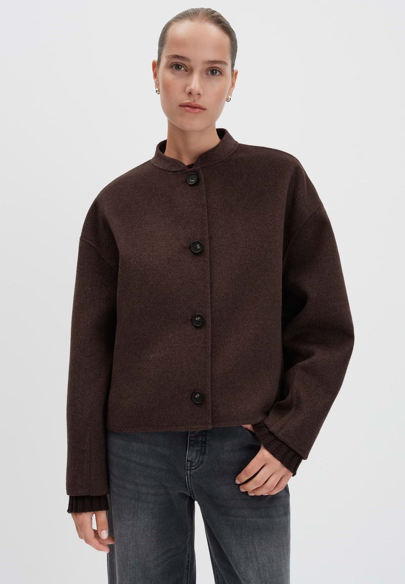 Vrouw in een donkerbruine gewatteerde wollen jas met een ronde kraag en donkergrijze jeans, staand tegen een effen lichte achtergrond.