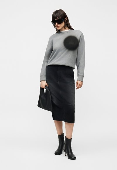 Frau trägt grauen Sweatshirt mit langen Ärmeln, schwarzen Midirock, schwarze Stiefeletten, große schwarze Sonnenbrille und hält schwarze Handtasche vor weißem Hintergrund.