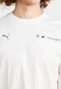 Camiseta de algodón blanca con un logotipo gris de Puma y la marca BMW Motorsport. Incluye el emblema de BMW y franjas rojas/azules en el frente.