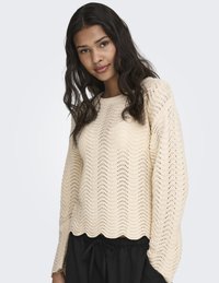 Jeune femme aux cheveux longs et ondulés portant un pull en maille beige avec des motifs ondulés et un ourlet festonné, associé à un pantalon noir à cordon de serrage.