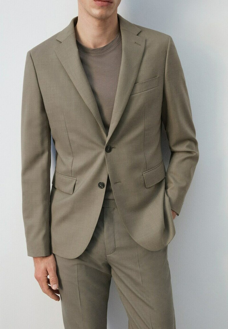 Man draagt een beige, op maat gemaakt pak met een enkelrijig jasje en bijpassende broek, gecombineerd met een taupekleurig shirt met ronde hals.