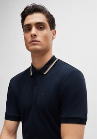 Camisa polo de color azul marino con mangas cortas, que presenta un cuello a rayas en beige y dorado, y un logo sutil en el pecho. Textura suave.