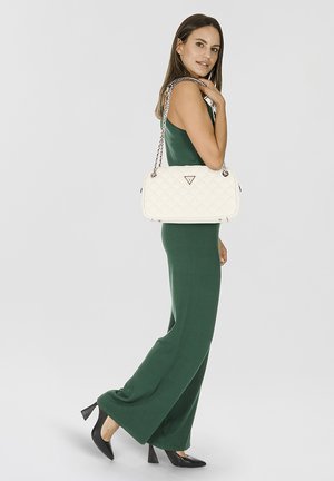 Witte quilted handtas met een kettingriem, voorzien van een driehoekig logo. Model draagt een groene jumpsuit en zwarte hoge hakken.