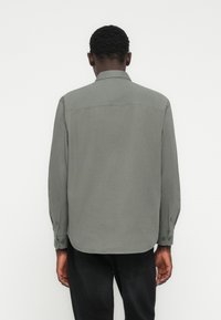 Γκρι ανοιχτό πουκάμισο με γιακά, μακριά μανίκια και ορατές ραφές· λεία υφή, εφαρμοστό στη μέση, φορεμένο με μαύρο παντελόνι.