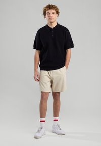 Schwarz geripptes Poloshirt mit kurzen Ärmeln, kombiniert mit beigen Shorts. Weiße Sneakers mit rot gestreiften Socken. Einfarbiger grauer Hintergrund.