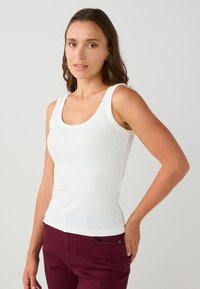 Weißes, geripptes Tanktop mit Rundhalsausschnitt und breiten Trägern, körperbetontes Design, kombiniert mit burgunderfarbenen Hosen mit seitlichen Taschen.