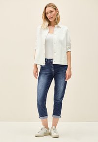 Veste légère blanche boutonnée par-dessus un débardeur blanc, associée à un jean en denim bleu foncé retroussé et des baskets blanches.
