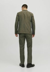 Két részes öltöny textúrált, sötétzöld anyagból. A zakó álló hajtókával rendelkezik, a slim-fit nadrágok pedig szabottak. Fekete cipőt visel.