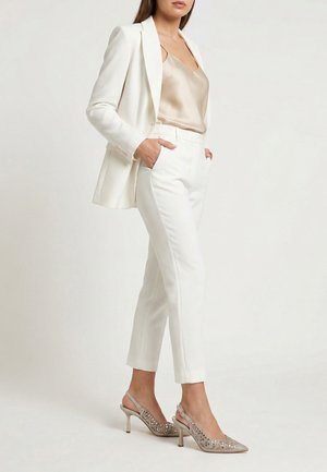 Frau trägt weiße maßgeschneiderte Hose und Blazer mit beiger Satin-Camisole und verzierten High-Heel-Slingback-Schuhen, Hände in den Taschen.