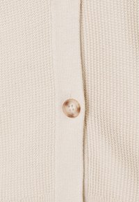 Chaqueta de punto color crema con un patrón texturizado, cierre con un botón marrón claro y bordes acanalados a lo largo de la tapeta.