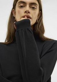 Haut noir à manches longues avec un col montant, présentant des poignets côtelés et une texture lisse. La main du modèle repose près du visage.