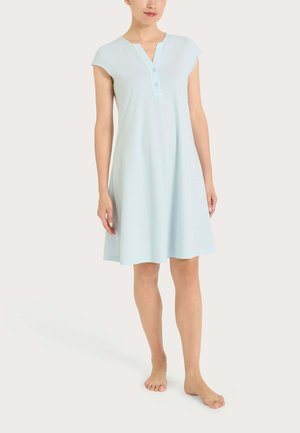 Donna in piedi a piedi nudi che indossa un vestito azzurro chiaro a maniche corte, lungo fino al ginocchio, con colletto a V abbottonato.