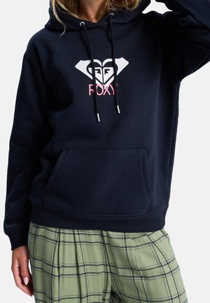 Osoba nosząca czarną bluzę z kapturem z białym logo w kształcie serca i różowym napisem „FOXY”, w połączeniu z zielonymi spodniami w kratę.
