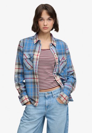Jeune femme aux cheveux courts bruns portant une chemise à carreaux bleue par-dessus un crop top rayé bordeaux et blanc ainsi qu’un jean bleu clair, les mains dans les poches.