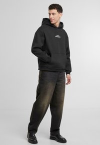 Sudadera negra con bolsillo frontal, con el texto blanco "piensa DIFERENTE". Corte relajado, combinada con unos pantalones vaqueros oscuros y deslavados de gran tamaño y zapatos negros.