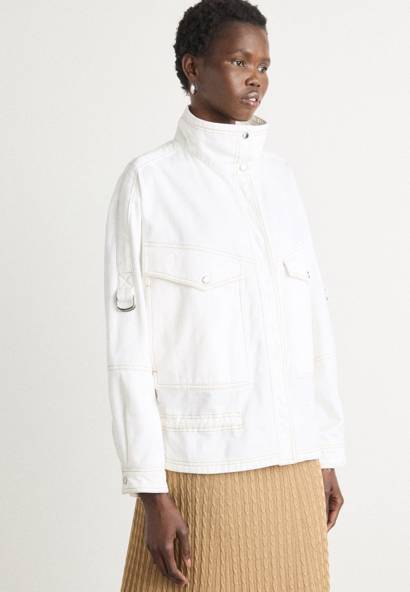 White denim jacket with a high collar, two front pockets, and snap closures; featured stitching in a lighter shade; worn over a ribbed beige skirt.