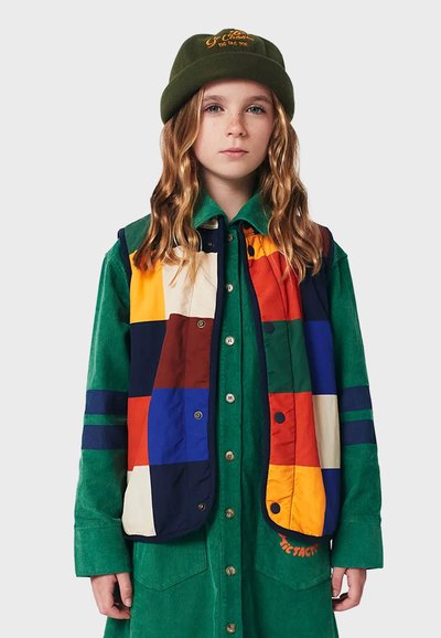 Bunt gemusterte Weste mit navyblauen, roten, gelben und grünen Abschnitten; kombiniert mit einem grünen Cordkleid mit Knopfverschluss.