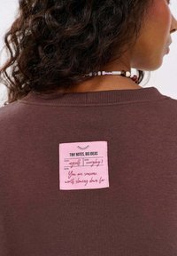 Hnědá mikina s růžovou etikou s ručně psanými poznámkami, umístěná na zadní straně. Měkká tkanina s volným střihem a žebrovaným výstřihem.