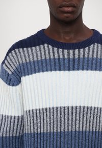 Homme portant un pull en tricot côtelé avec des rayures horizontales dans des tons marine, gris, bleu, blanc et charbon, visible des épaules aux lèvres.