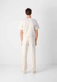 Man with short brown hair wearing a cream t-shirt, cream straight-leg pants, and beige shoes, standing facing a white wall.