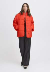 Kvinde med bølget brunt hår iført en lys orange quiltet jakke, sorte vide bukser og sorte sko, stående mod en ensfarvet baggrund.