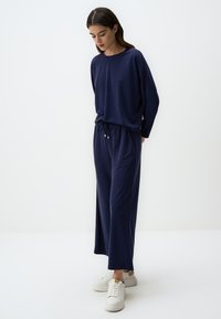 Marineblaues Langarmoberteil mit lockerer Passform und elastischem Taillenbund, aus weichem Material gefertigt. Kombiniert mit weißen Sneakers und minimalem Schmuck.