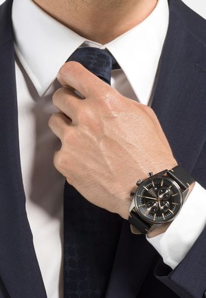 Black chronograph watch with a textured black strap, silver accents, and a multi-dial face, worn on a hand adjusting a blue patterned tie.