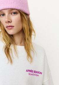 Biała bluza z fioletowym haftem o treści „APRÈS SEASON Beyond the Slopes”. Modelka nosi różową dzianą czapkę, prezentując długie, falowane włosy.