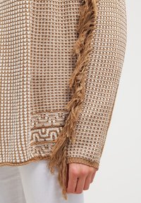 Sweter dzianinowy w kolorach beżowym i kremowym, z teksturowanym wzorem, frędzlami po bokach oraz geometrycznym detalem na końcu.