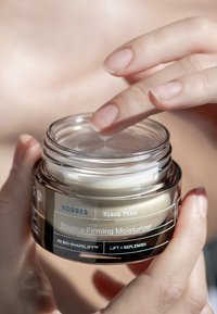 En genomskinlig glasburk med Korres Black Pine Bounce Firming Moisturizer, krämfärgad produkt inuti, hålls av händer med en naturlig finish.