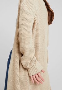 Beżowy sweter o luźnym kroju, z prążkowanymi mankietami i detalami. Gruba tekstura z widocznymi szwami, otwarty przód.