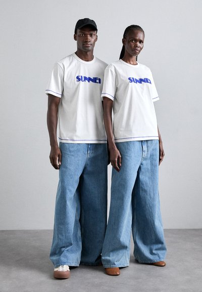 Λευκά T-shirts με μπλε κείμενο "SUNNEI", συνδυασμένα με χαλαρές τζιν παντελόνες ανοιχτού τόνου. Καθημερινά υποδήματα, μινιμαλιστικός σχεδιασμός και απλές λεπτομέρειες.