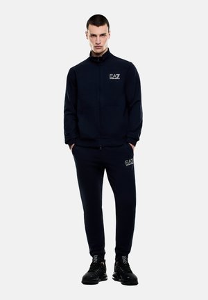 Hombre de pie con las manos en los bolsillos, vistiendo chándal negro EA7 Emporio Armani y zapatillas negras sobre fondo liso.