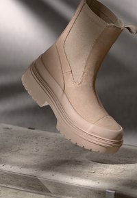 Botín beige con un panel de malla texturizado, suela gruesa y puntera reforzada. Diseño ligeramente estructurado con detalles de costura mínimos.