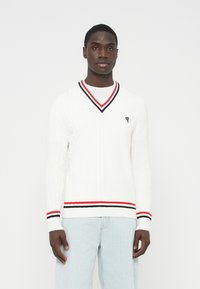 Baltas megztas megztinis su V formos iškirpte, apykaklėje ir apačioje raudonos bei juodos spalvų juostos. Ant krūtinės mažas juodas širdies logotipas.