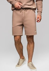 Shorts en coton marron avec une taille élastique et un cordon de serrage. Poignets côtelés à l'ourlet, associés à des baskets beiges.