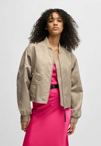 Beige lyhyt bomber-takki, jossa on sileä pinta, vetoketjukiinnitys ja joustavat hihansuut, kerroksittain vaaleanpunaisen satiinimekon päällä.