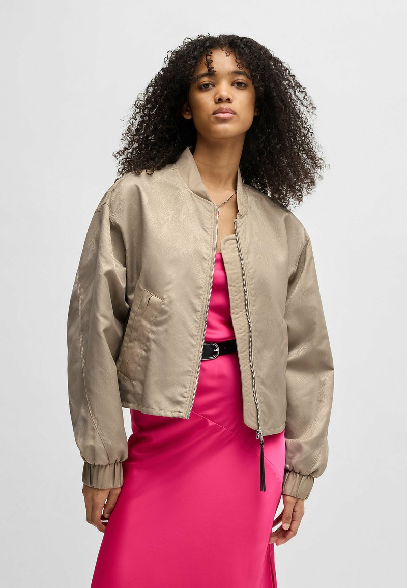 Beige lyhyt bomber-takki, jossa on sileä pinta, vetoketjukiinnitys ja joustavat hihansuut, kerroksittain vaaleanpunaisen satiinimekon päällä.