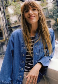 Džínová bunda so striebornými gombíkmi, oblečená cez čierno-biele pruhované tričko. Dlhé vlasy padajú cez plecia na rozmazanom vonkajšom pozadí.