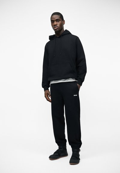 Homme portant un sweat à capuche noir, un pantalon de survêtement noir avec le logo blanc "MSGM" et des baskets noires, debout devant un fond blanc.