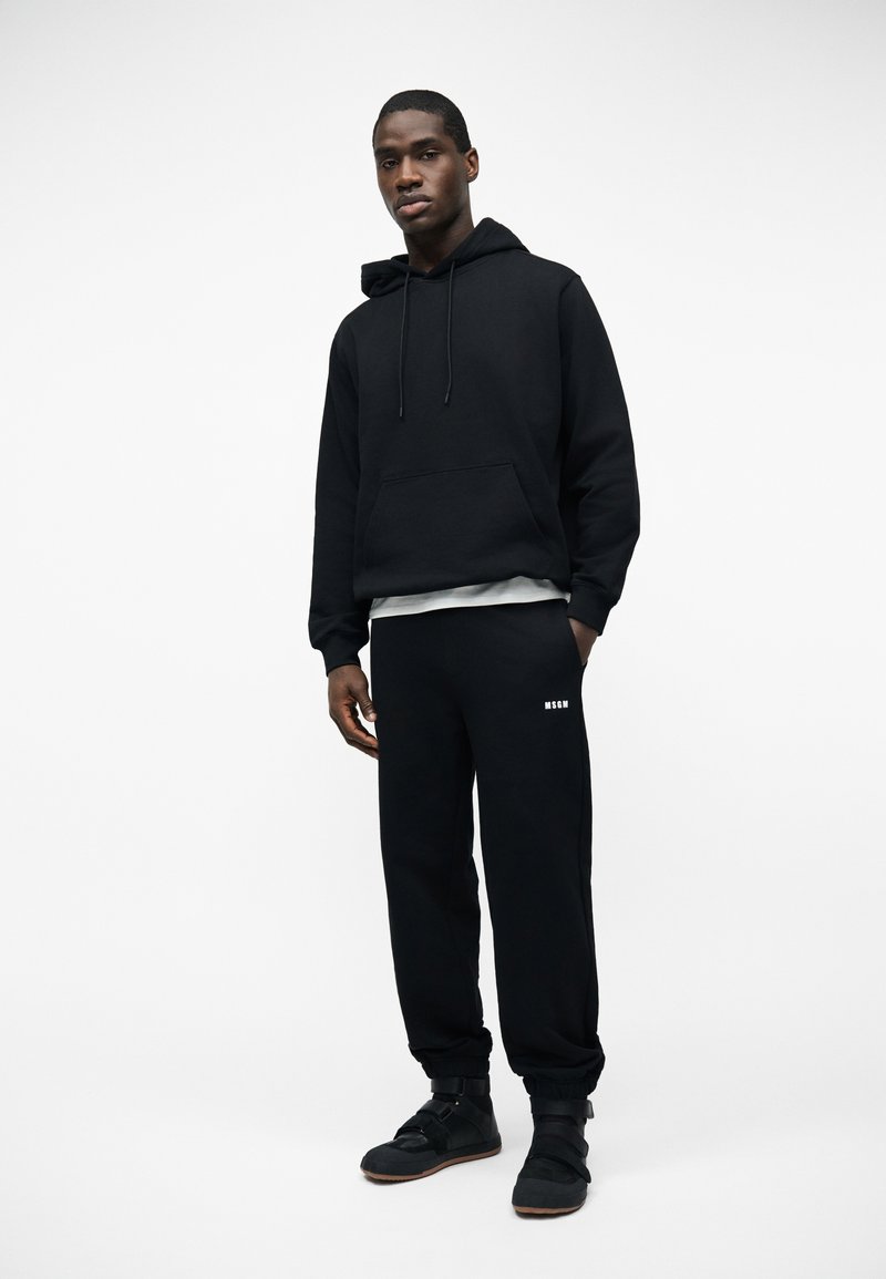 Man in een zwarte sweat met capuchon, zwarte joggingbroek met wit "MSGM"-logo en zwarte sneakers, staand tegen een witte achtergrond.
